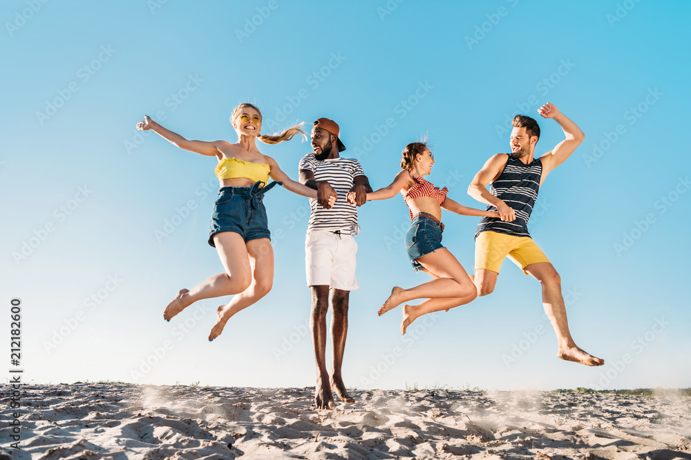 happy young multiethnic friends holding hands and jumping in sandy beach