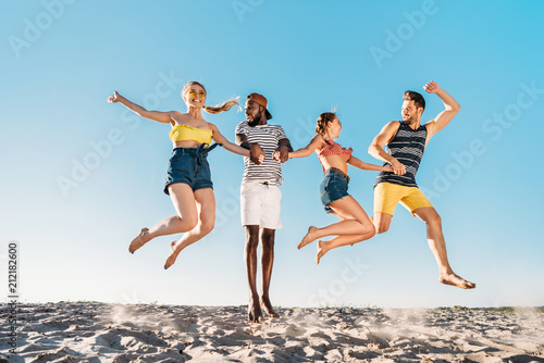 happy young multiethnic friends holding hands and jumping in sandy beach