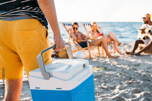 cropped shot of man holding beach cooler while friends resting on sand behind photo