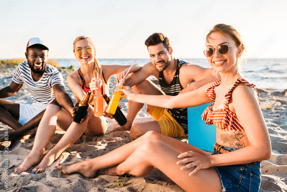 happy young multiethnic friends holding glass bottles with drinks and smiling at camera on sandy beach