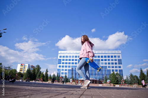 A beautiful girl with a skateboard in her hands is walking on a background of city skyscrapers.