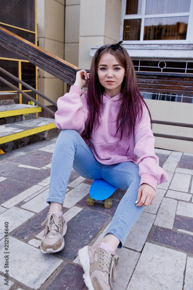 Fototapeta premium Cheerful young woman in sunglasses sitting on skateboard and having fun over pink background