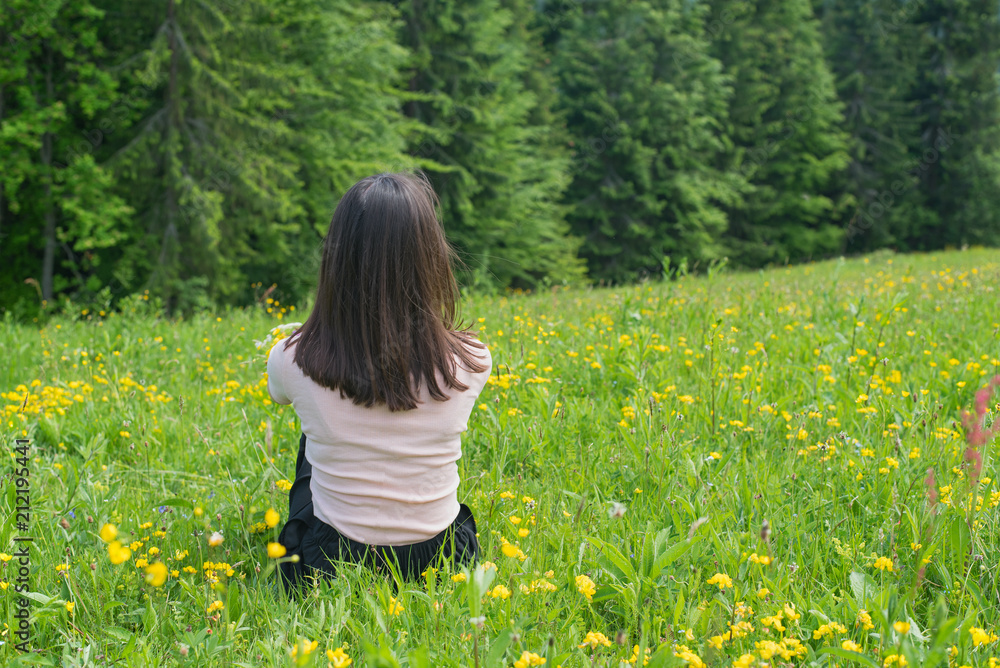 Fototapeta premium Girl sits on a green lawn near a forest. Back view
