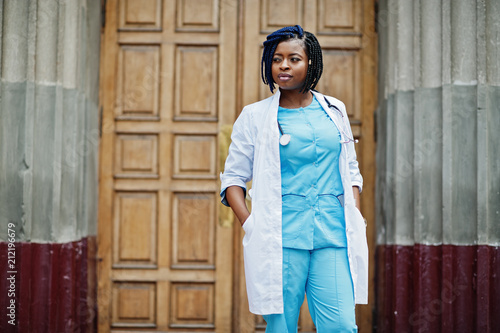 Stylish african american doctor with stethoscope and lab coat posed against door of hospital.