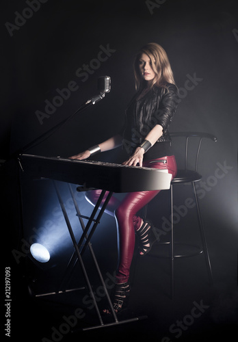 Blonde girl musician pianist plays perform on white digital piano, sits on chair, sings into retro microphone. Woman teacher shows how to play music on black background