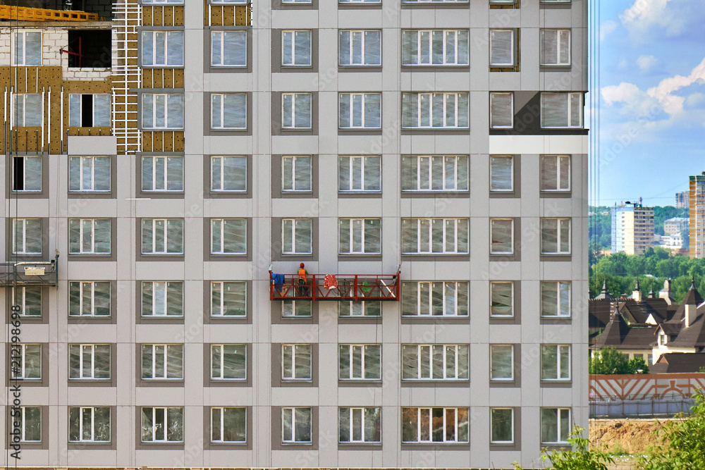 Worker in a construction cradle on the wall of a building under construction with a cityscape behind the building Worker in a construction cradle on the wall of a building under construction with a cityscape behind the building