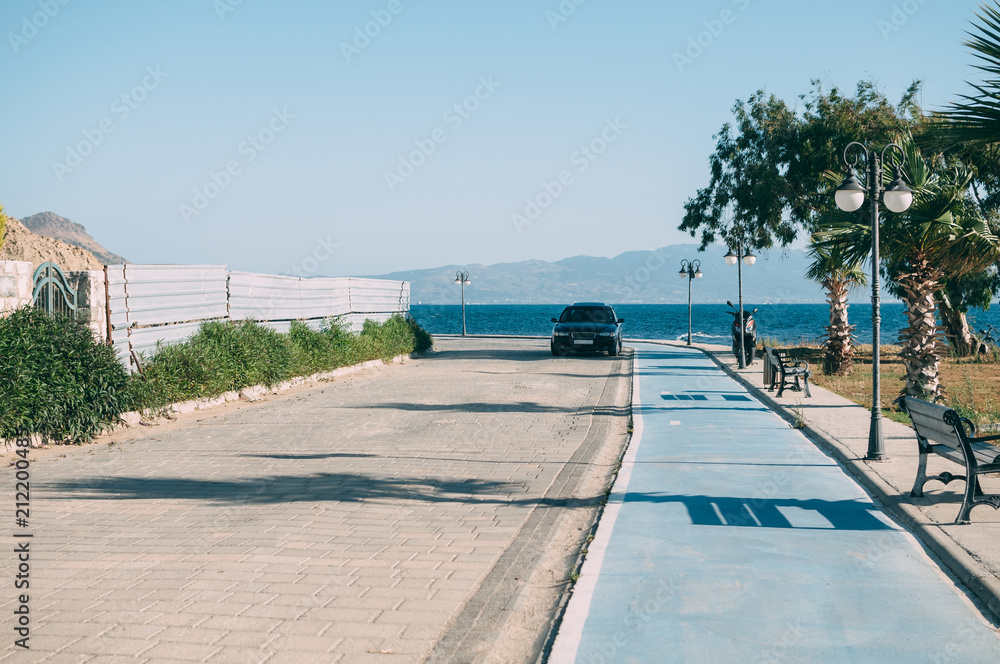 On the embankment in the shade of palm trees there is a black car on the background of the sea and mountains.