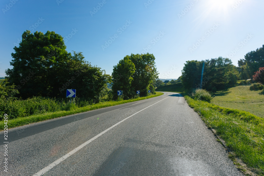 Empty asphalt country road passing through green fields and forest. Countryside landscape on sunny day with sunbeams in the blue sky in France. Transport, industrial agriculture, road network concept.