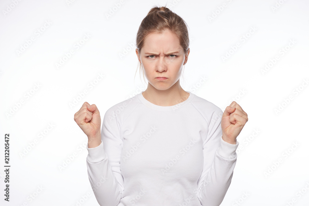 Offended European lady looking cute when angry. Portrait of outraged tender feminine woman, raising clenched fists and frowning, being insulted or mad, sulking from unhappiness over white background. Offended European lady looking cute when angry. Portrait of outraged tender feminine woman, raising clenched fists and frowning, being insulted or mad, sulking from unhappiness over white background.