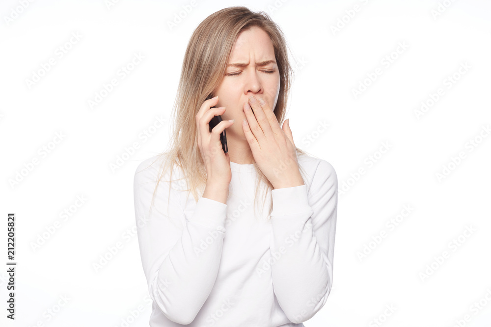 Indoor shot of tired and bored cute female in white t-shirt, yawning, covering opened mouth with palm, trying not to fall asleep during daytime, holding phone, overworking or hearing boring talks. Indoor shot of tired and bored cute female in white t-shirt, yawning, covering opened mouth with palm, trying not to fall asleep during daytime, holding phone, overworking or hearing boring talks.
