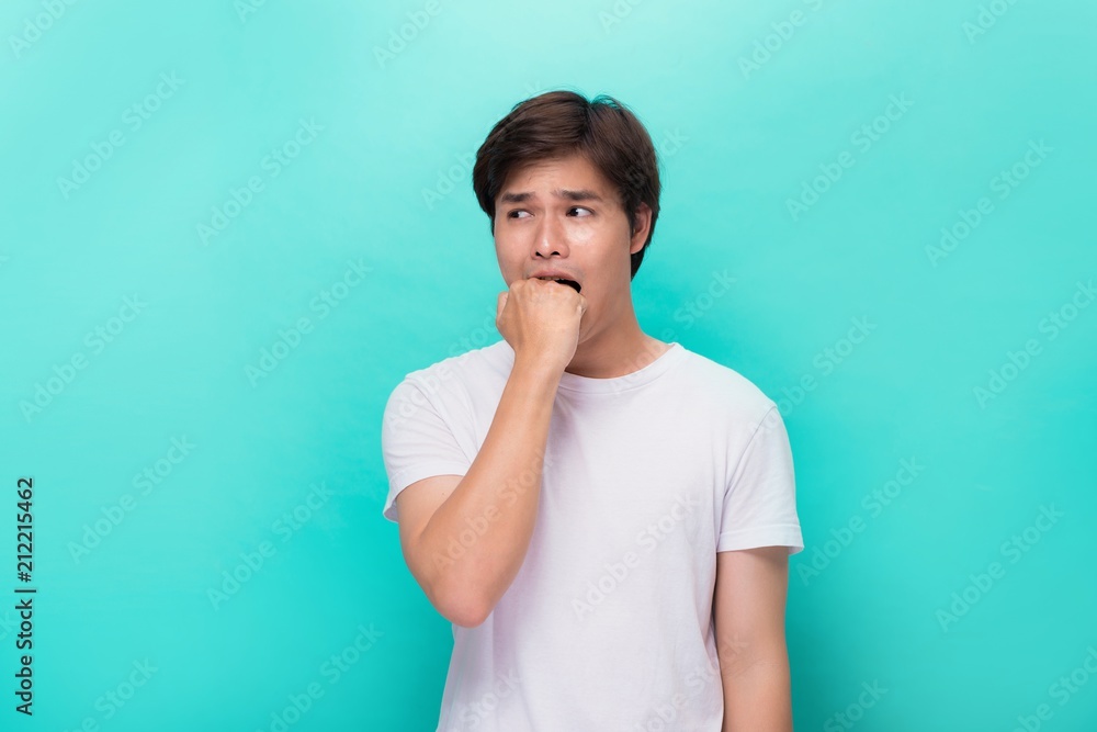 Angry irritated aggressive young bearded man in white t-shirt biting his fist over blue background Angry irritated aggressive young bearded man in white t-shirt biting his fist over blue background