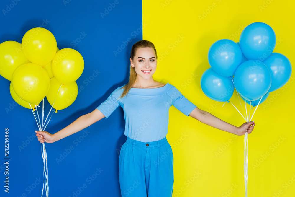 Fototapeta premium Smiling blonde woman holding blue and yellow balloons on blue and yellow background