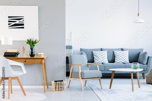 Real photo of a living room interior with study space. Typewriter on a wooden desk next to a sofa with two white cushions and houndstooth pattern armchair