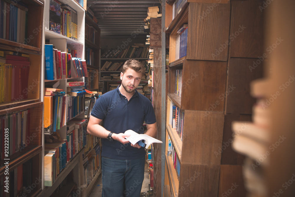 Man with a book in his hands stands in an atmospheric public library and looks at the camera. Portrait of student man with beard and black shirt in public library.