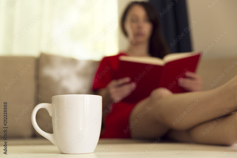 Woman with cup of hot tea reading book by window on sofa at home. Focus on cup of tea Woman with cup of hot tea reading book by window on sofa at home. Focus on cup of tea