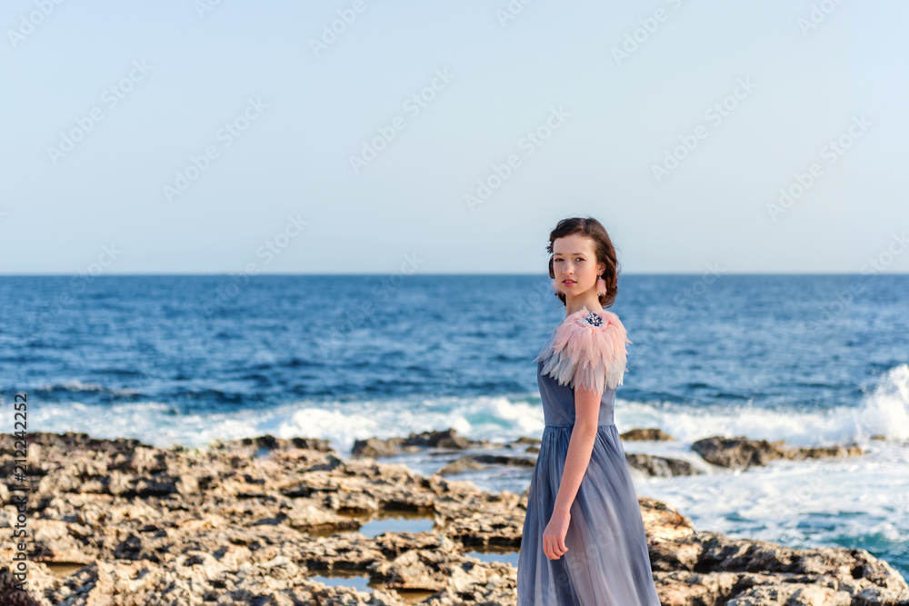 portrait of a young slender graceful girl in a gray dress with pink feathers with a raging bright blue sea on the background portrait of a young slender graceful girl in a gray dress with pink feathers with a raging bright blue sea on the background