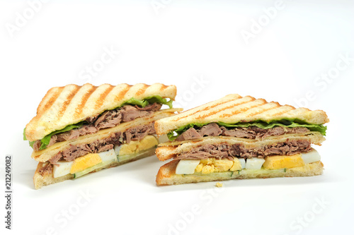 Two sandwiches of triangular shape made of white bread. The filling of the sandwich consists of pieces of tuna, hard boiled eggs, lettuce leaves, pieces of cheese and sauce. Closeup. White background.