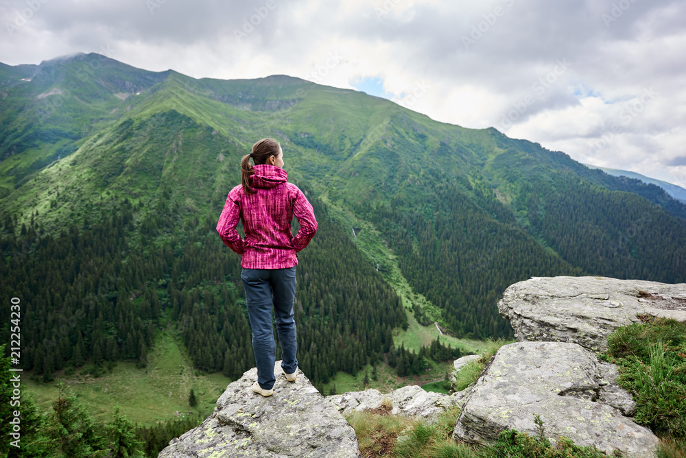 Rear view of a girl standing on a cliff in front of her opening a mesmerizing landscape of mighty green mountains and clouds above them. Europe, Romania
