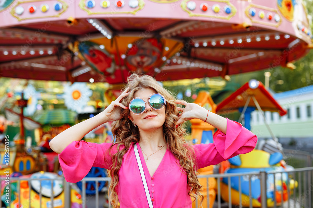 Bright summer concept. A cheerful blonde girl in pink jacket and stylish sunglasses is having fun in the amusement Park. Cheerful woman smiling against bright carousel