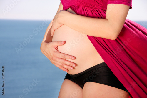 Beautiful pregnant woman and lady is hugging and holding her pregnant belly, standing in natural environment. Waiting for baby. Mom holds hands on belly on sea and sky nature background. Pregnancy.