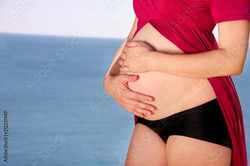 Beautiful pregnant woman and lady is hugging and holding her pregnant belly, standing in natural environment. Waiting for baby. Mom holds hands on belly on sea and sky nature background. Pregnancy.