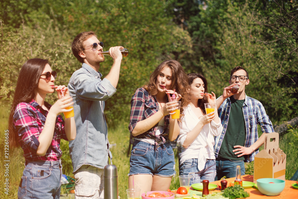 Fototapeta premium Five people outdoors on sunny day eating and drinking. Joyful company of young people out on picnic having drinks and food. Trees in background.