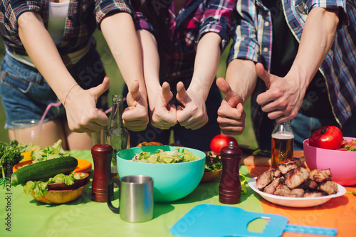 Group of friends showing thumbs up to table of food. Group of young peope showing thumbs up signs with both hands to big table of picnic food they have prepared. photo