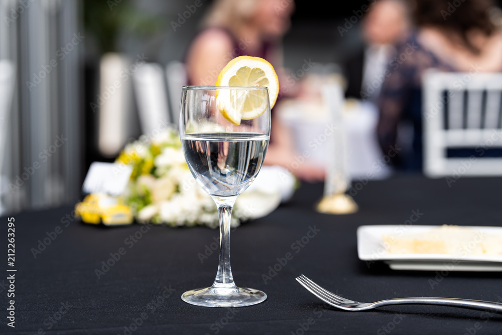 Closeup of one glass with lemon slice and water in restaurant or wedding reception black table tablecloth, bokeh of people, plate with cake slice, bouquet flowers, fork, chairs, dishes
