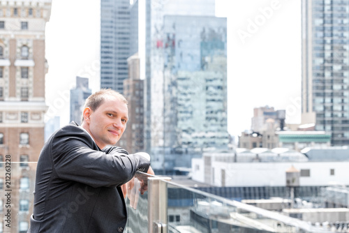 Young businessman portrait standing in suit, tie, looking at New York City cityscape skyline in Manhattan at skyscrapers rooftop happy smiling © Kristina Blokhin
