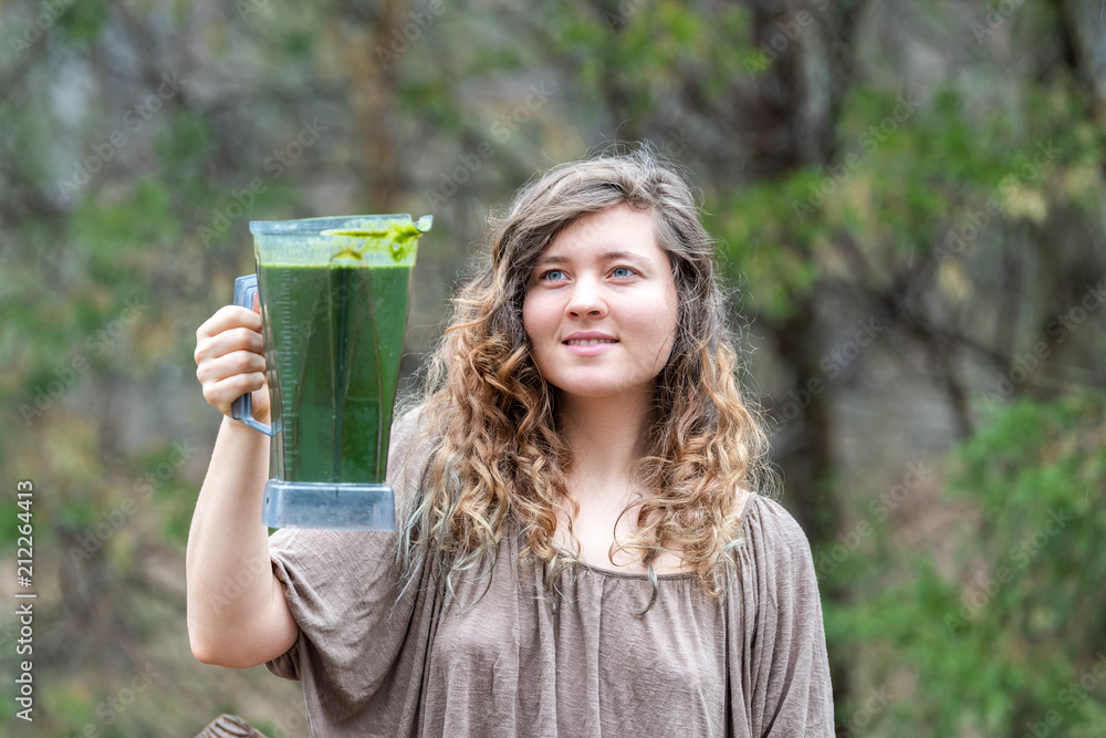 Young woman outside, outdoors, holding plastic blender container for smoothie with green vegetables Young woman outside, outdoors, holding plastic blender container for smoothie with green vegetables