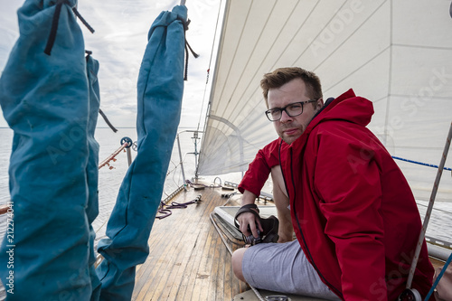A man - a photographer in a red jacket and glasses sits on the deck of a moving yacht and holds a camera in the hands  against the background of the sea and sky in the clouds  on a summer day.