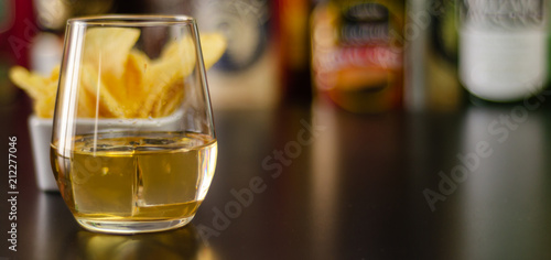 glass of whiskey with ice cubes and salty snacks on the background of bottles and bar