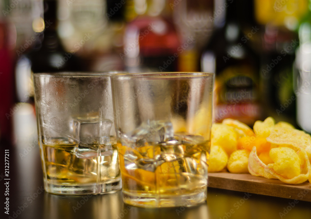 glass of whiskey with ice cubes and salty snacks on the background of bottles and bar