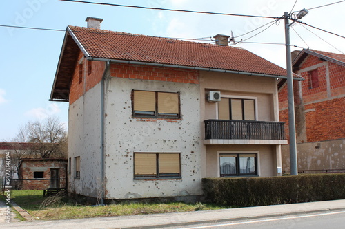 Partially renovated suburban house damaged by shrapnel during war with falling facade, damaged window frames, uncut grass, hedge, air condition unit and electrical pole in front