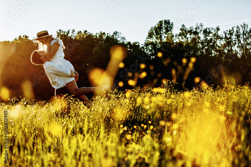 handsome guy with a beard in a white denim shirt gentle hugs, hand holding and kissing a girl with blond hair in a white dress and hat in a field at sunset.