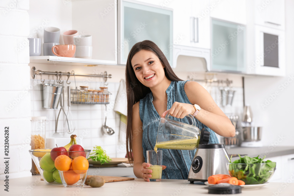 Young woman pouring tasty healthy smoothie into glass at table in kitchen