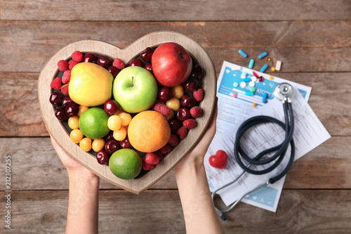 Female doctor holding plate with fresh fruits over wooden table, top view. Cardiac diet photo