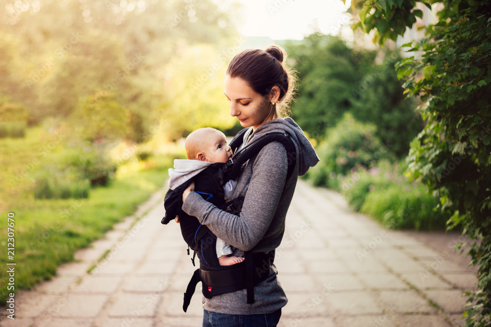 Little baby girl and her mother walking outside during sunset. Mother is holding and tickling her baby, babywearing in the ergo carrier Little baby girl and her mother walking outside during sunset. Mother is holding and tickling her baby, babywearing in the ergo carrier