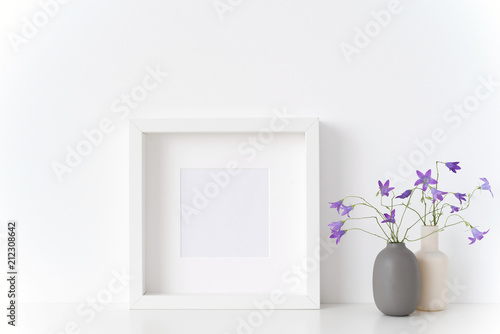 White square portrait frame mockup with spring bellflowers in vases near white wall on white background. Empty frame mock up for presentation design. Template framing for modern art. photo