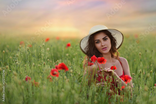 Beautiful young woman in red dress and white hat walks around field with poppies in a beautiful summer evening