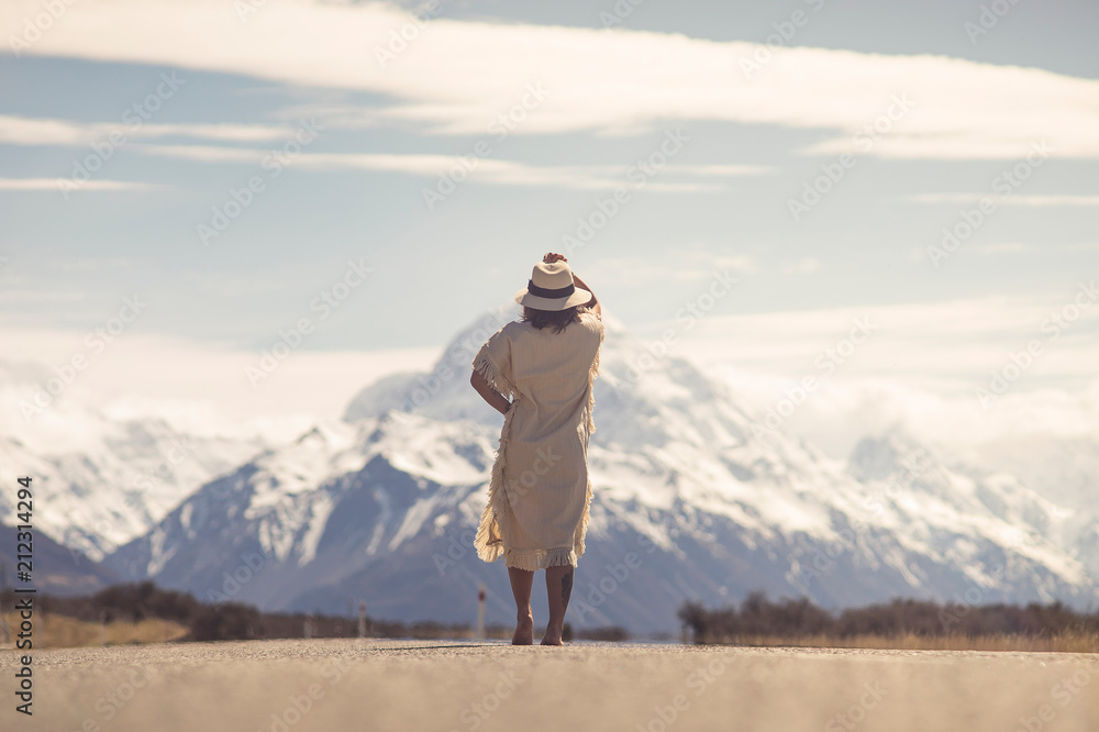 A boho hippy lady in white dress with hat stands in the middle on road with aoraki snow mountain background in New Zealand. idea for travel, destination.