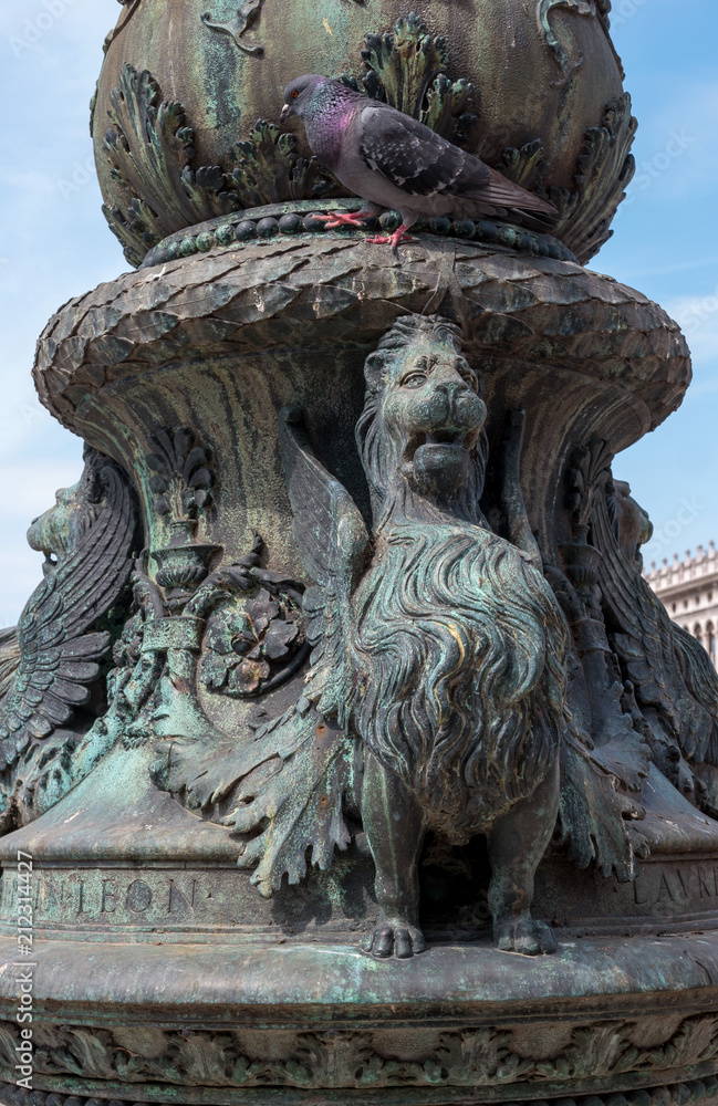 The Venetian winged lion in the decor of the support of a street lamp located next to Soboroma San Marco, the Doge's Palace and the Campanile. Pigeon walking on the lamp support. Venice, Italy
