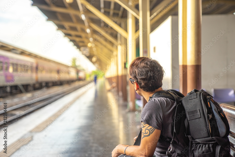 a traveller, backpacker man in casual clothes and sunglasses with a camera, sitting and waiting for a train at platform train station a traveller, backpacker man in casual clothes and sunglasses with a camera, sitting and waiting for a train at platform train station