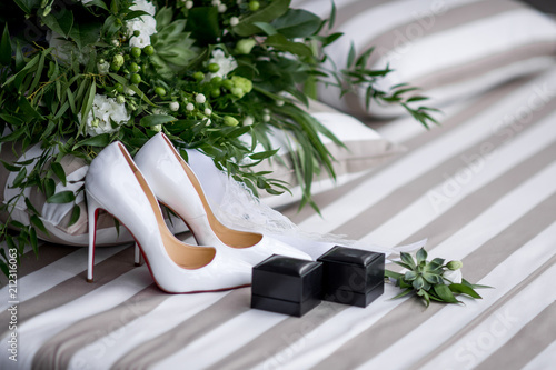 Golden wedding rings, white shoes and green bouquet stand on a dinner table
