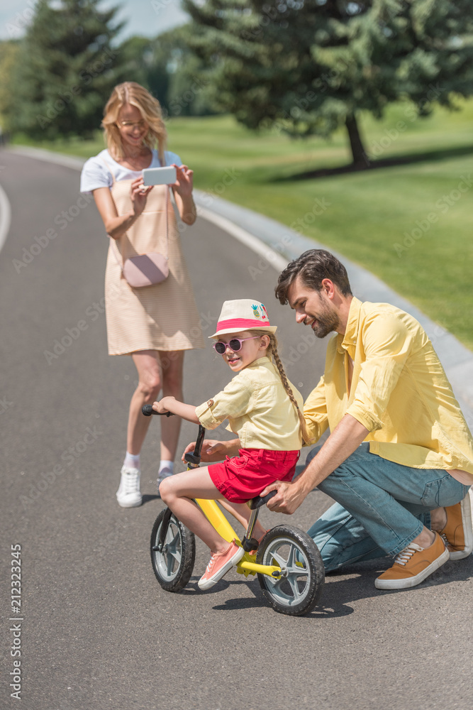 smiling young woman with smartphone photographing happy father teaching daughter riding bicycle smiling young woman with smartphone photographing happy father teaching daughter riding bicycle
