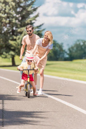 happy young parents looking at little daughter riding bicycle in park