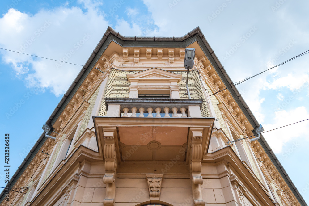 Corner of a historic 19th century building with balcony seen from the bottom up with blue sky and clouds in the background in Szeged, Hungary Corner of a historic 19th century building with balcony seen from the bottom up with blue sky and clouds in the background in Szeged, Hungary