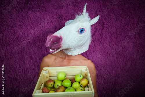 Fototapete Freaky young woman in comical mask stands on the purple background and shows box of apples