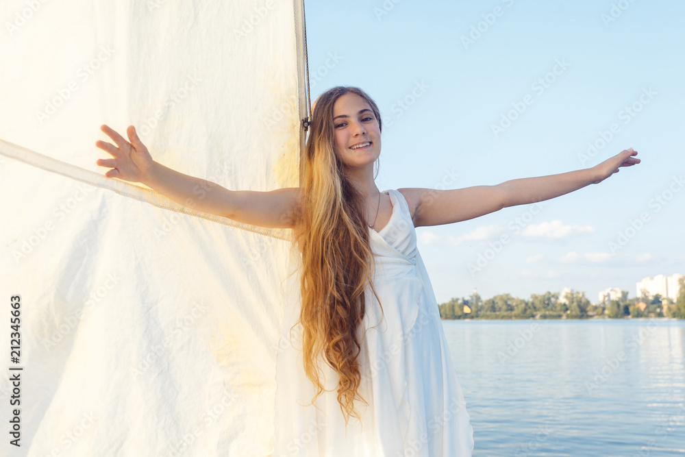 portrait of happy pretty girl with white dress and long curly blonde hair standing on yacht at summertime. standing near white sail with raised hands and looking at camera with toothy smile portrait of happy pretty girl with white dress and long curly blonde hair standing on yacht at summertime. standing near white sail with raised hands and looking at camera with toothy smile