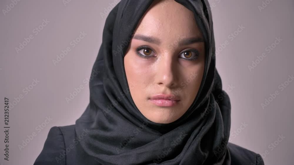 Young sad muslim girl in hijab is watching down and at camera, grey background, religious concept Young sad muslim girl in hijab is watching down and at camera, grey background, religious concept